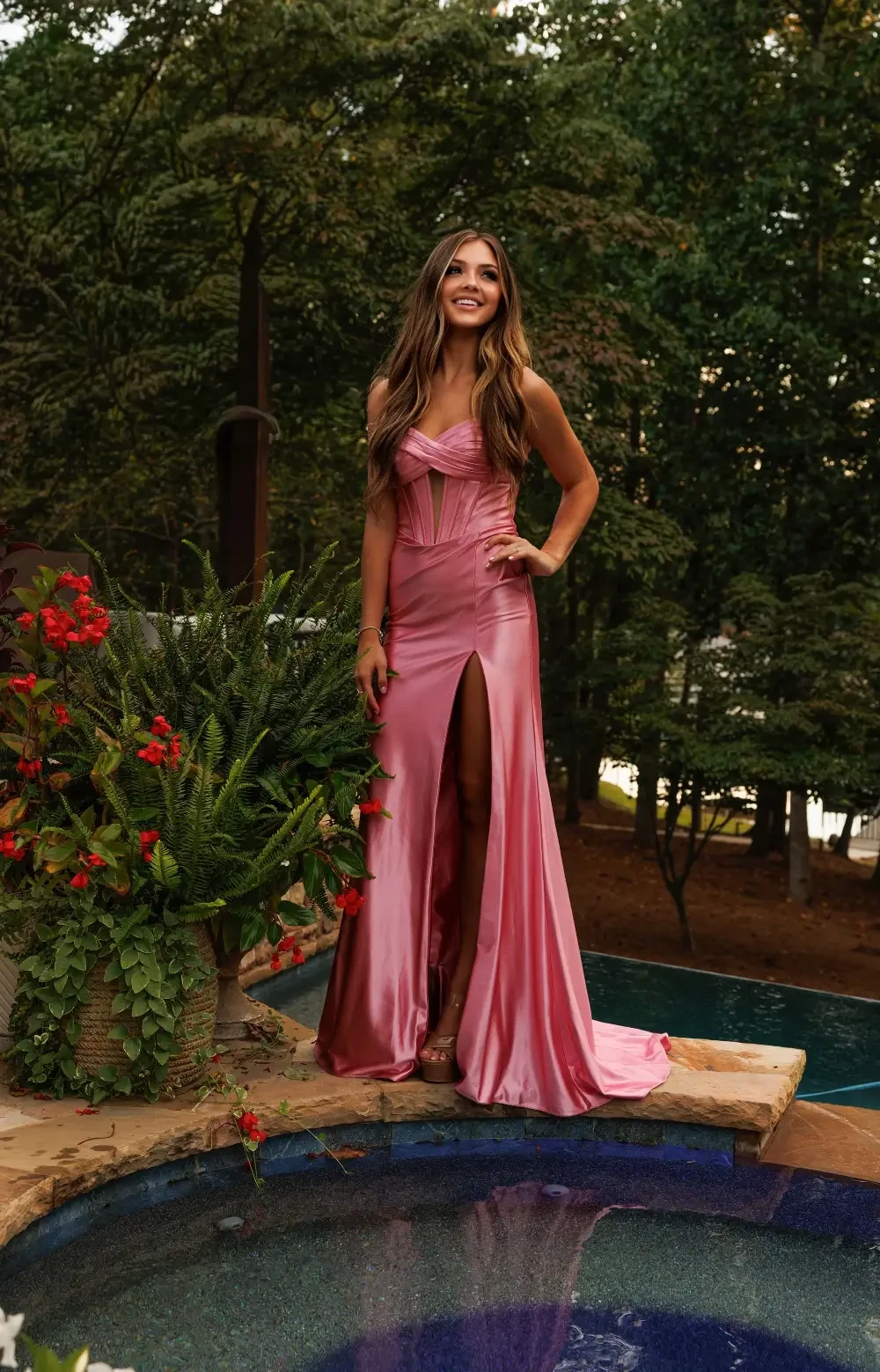 A woman in a pink dress standing by a pool surrounded by greenery and flowers.