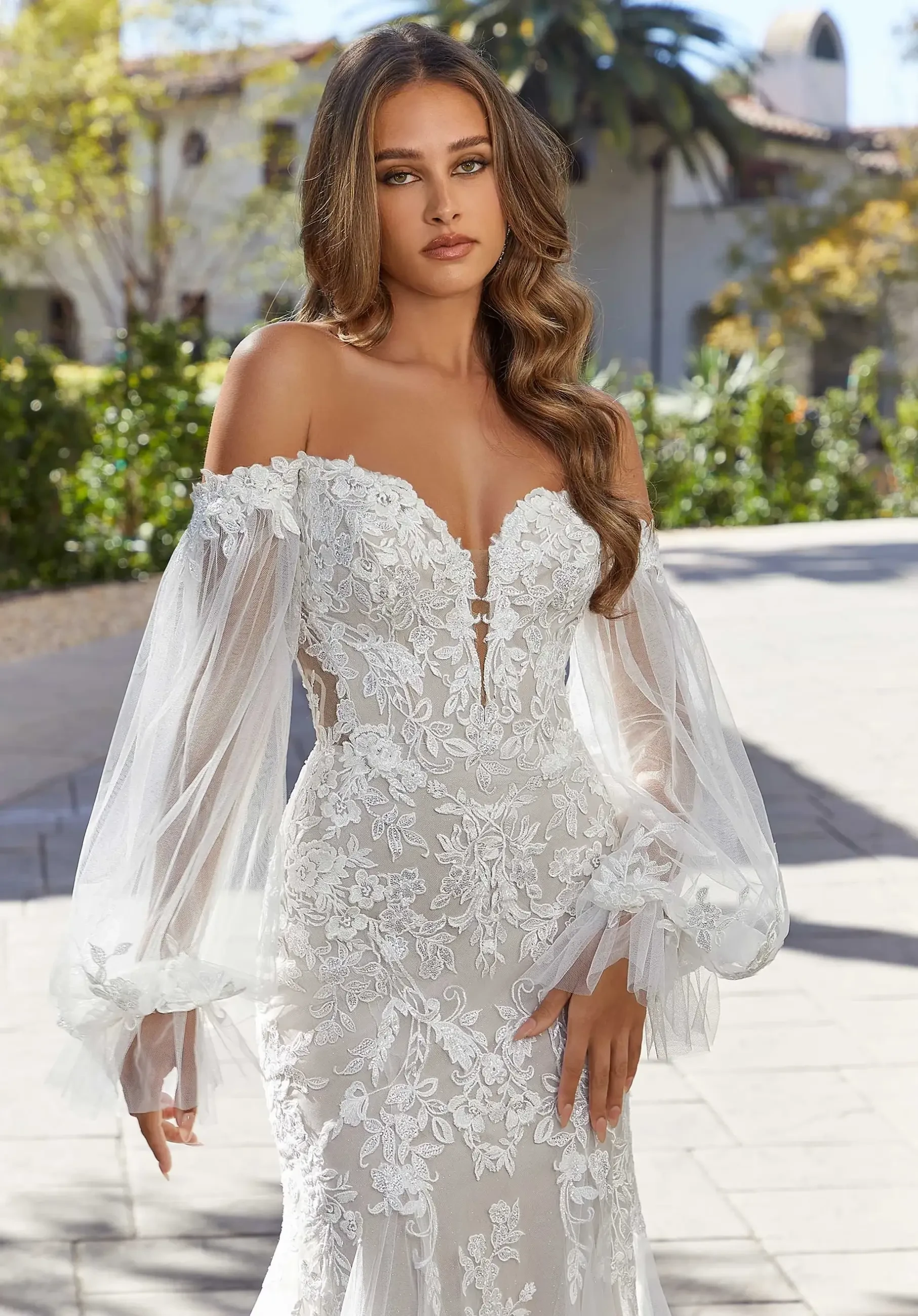 Model is wearing a sexy fit and flare gown with a plunging sweetheart bodice, intricate frosted, embroidered lace, an illusion back, and detachable whimsical bishop sleeves.