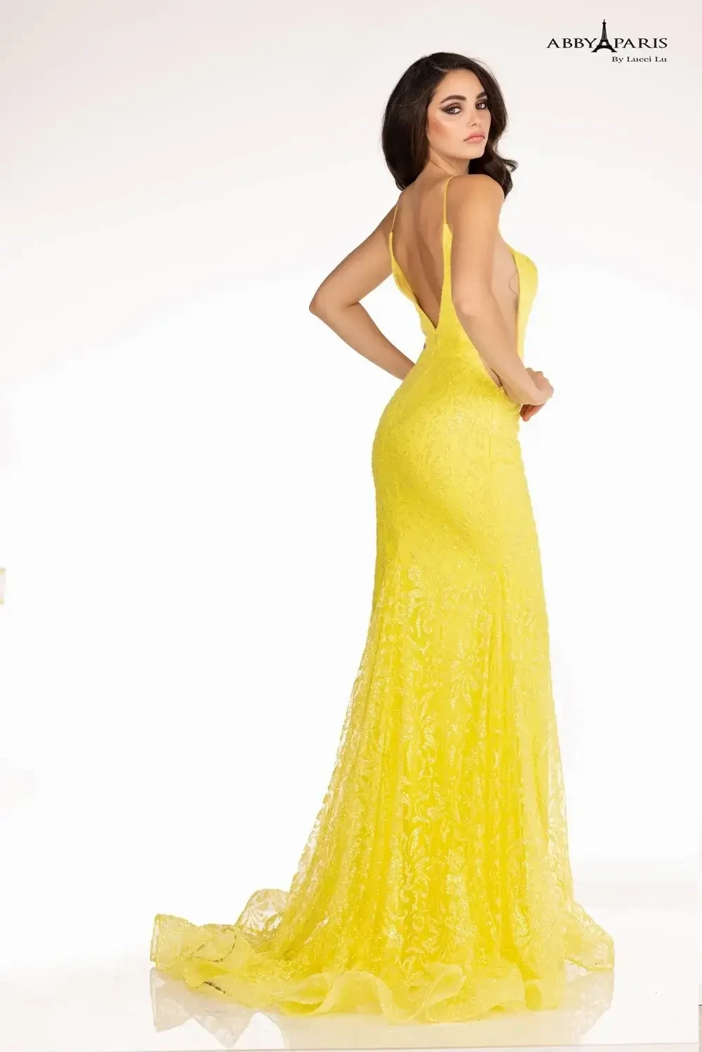 Woman in a vibrant yellow lace gown poses confidently against a white background. The dress features a low back and elegant train, conveying sophistication.