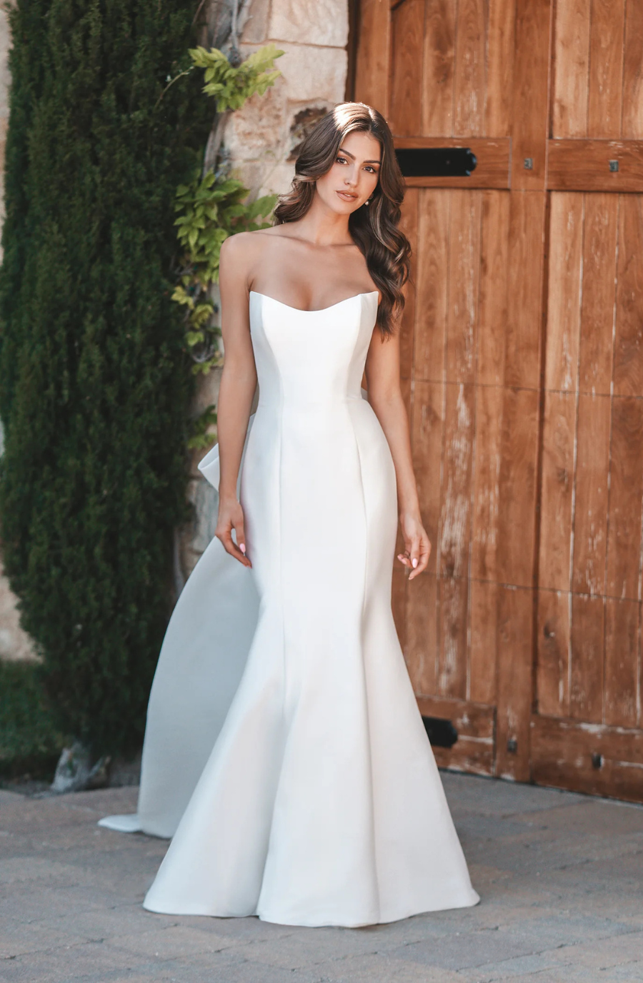 Woman in a sleek, strapless white gown stands by a wooden door and ivy-covered stone wall, exuding elegance and sophistication.