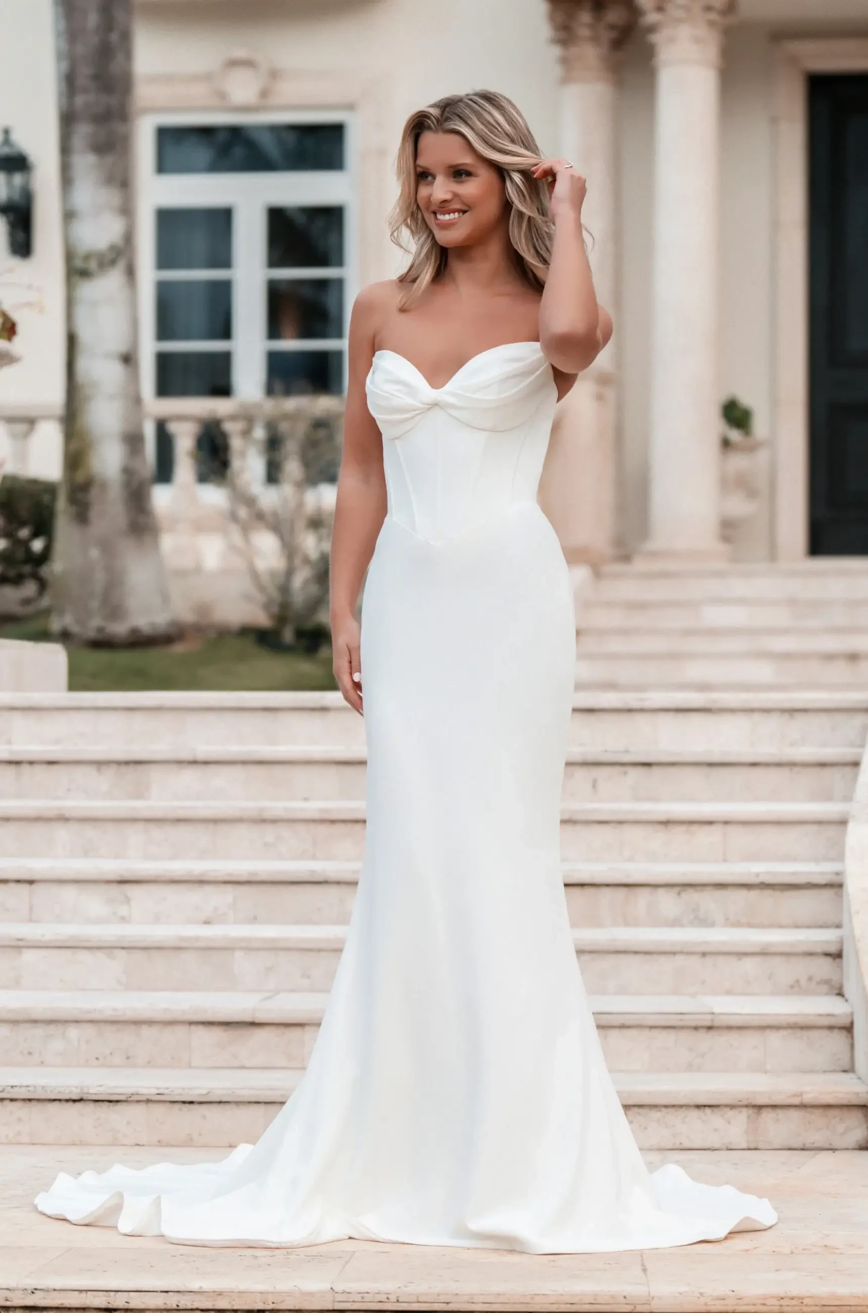 Model is wearing a sleek crepe sheath gown with a corset-style bodice, a draped sweetheart neckline, and detachable off-the-shoulder sleeves.