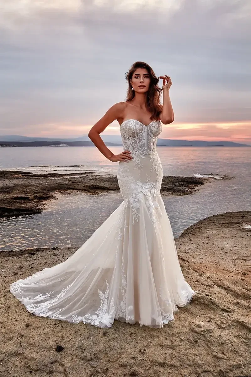 A woman wearing a strapless wedding gown stands on a rocky shoreline during sunset, with the water in the background.