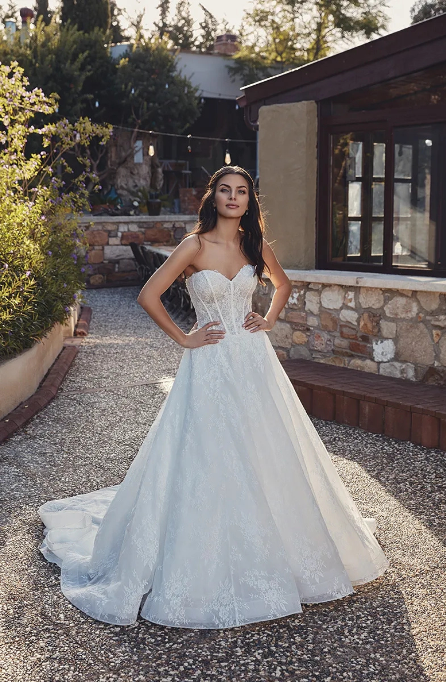 Bride in an elegant, strapless wedding gown stands in a sunlit garden with stone paths and lush greenery, exuding grace and confidence.