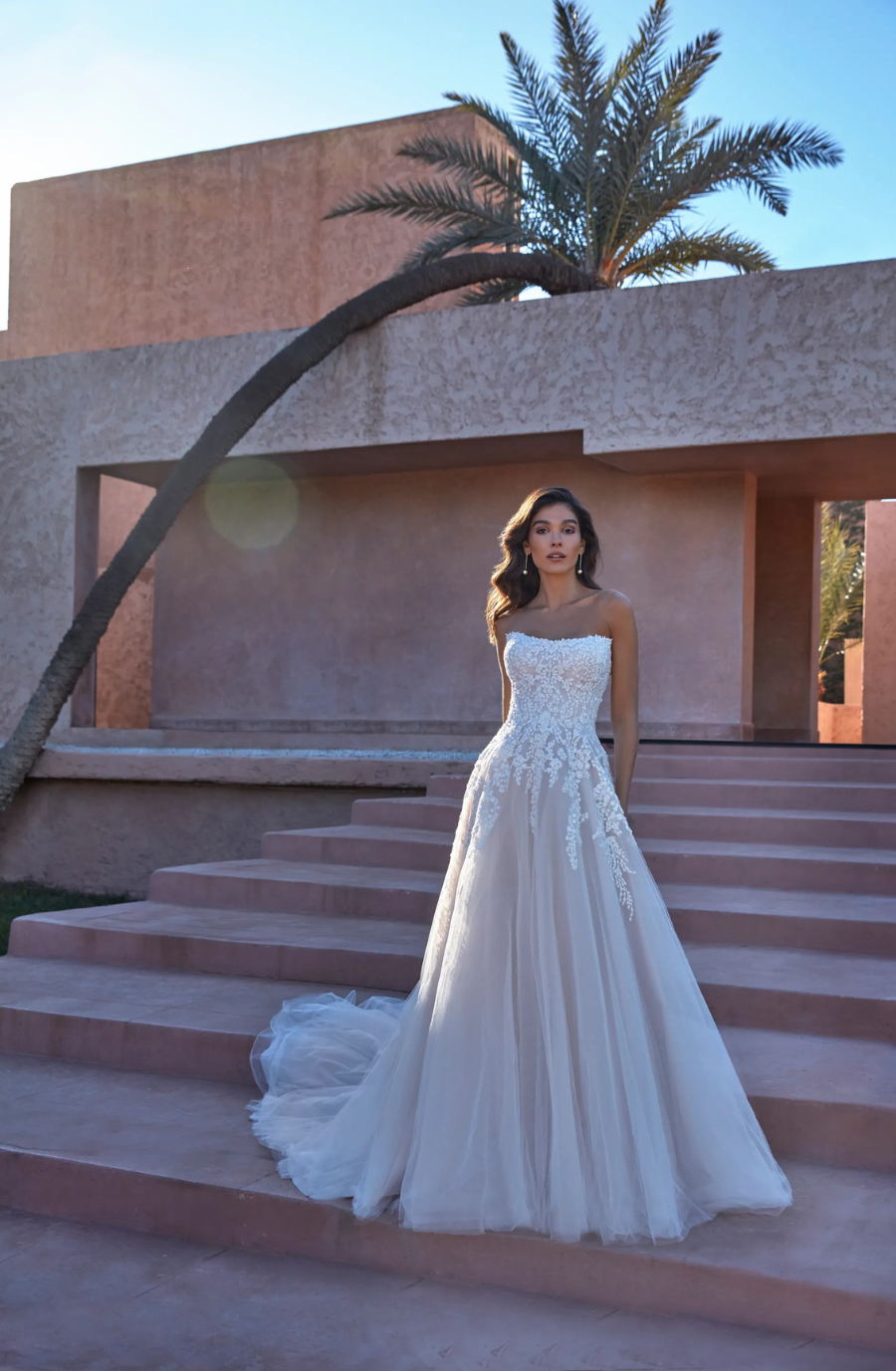 A woman in a strapless, lace wedding gown stands on pink stone steps with a palm tree in the background. The setting is serene and sunlit.