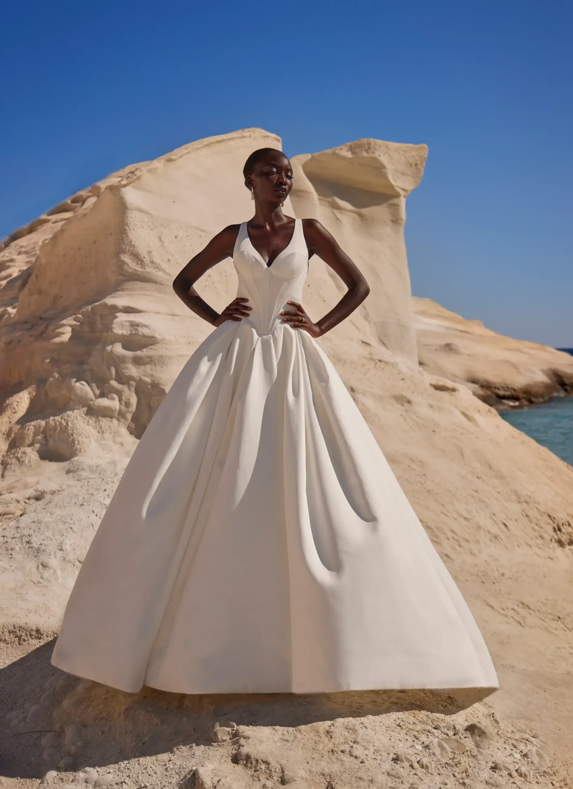 A woman wearing a white wedding dress stands confidently on a sandy beach, with rocky formations and the ocean in the background under a clear blue sky.