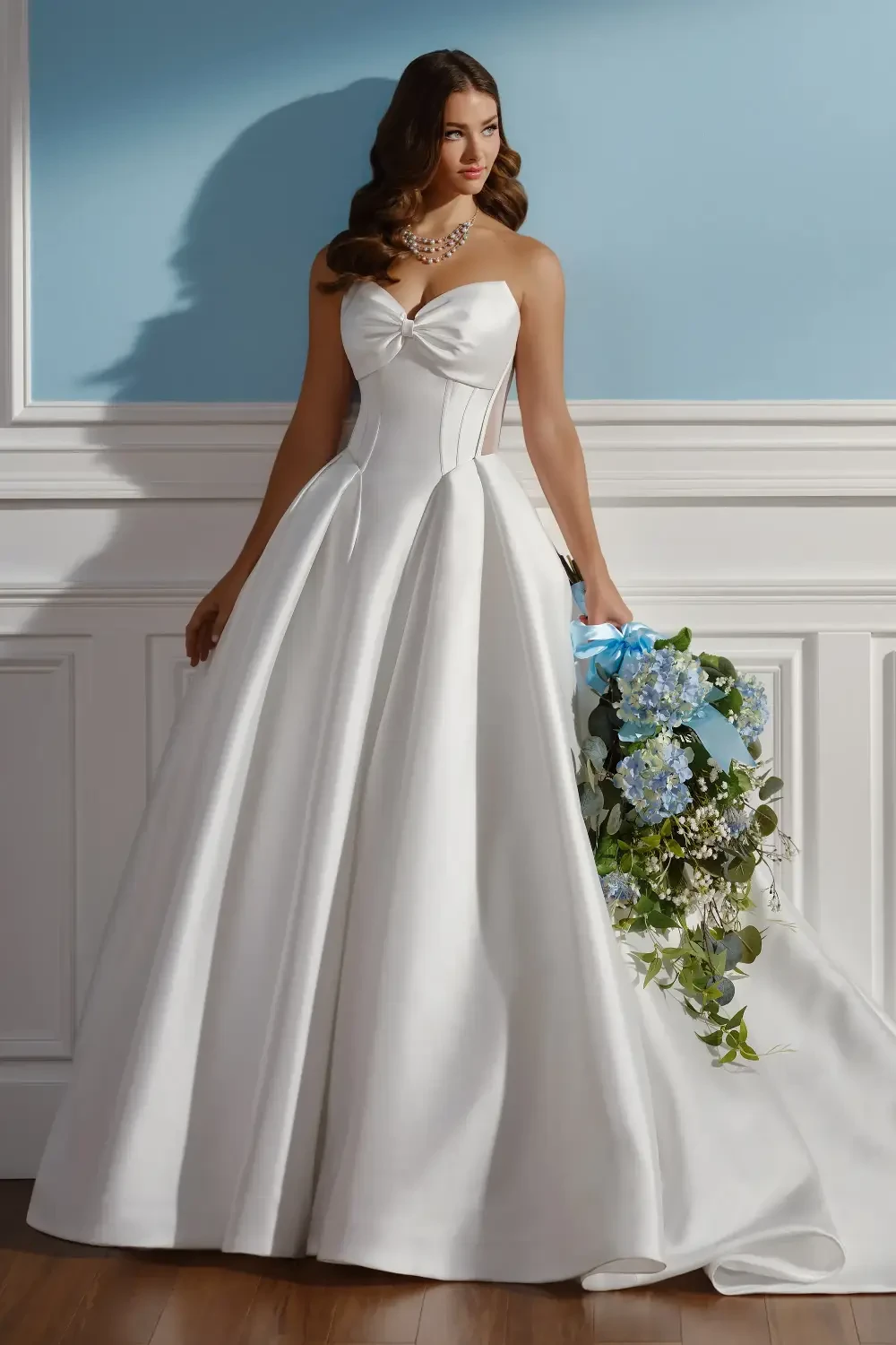 A woman wearing a strapless white wedding gown with a flowing skirt, holding a bouquet of blue flowers, standing in a bright room with a blue wall.