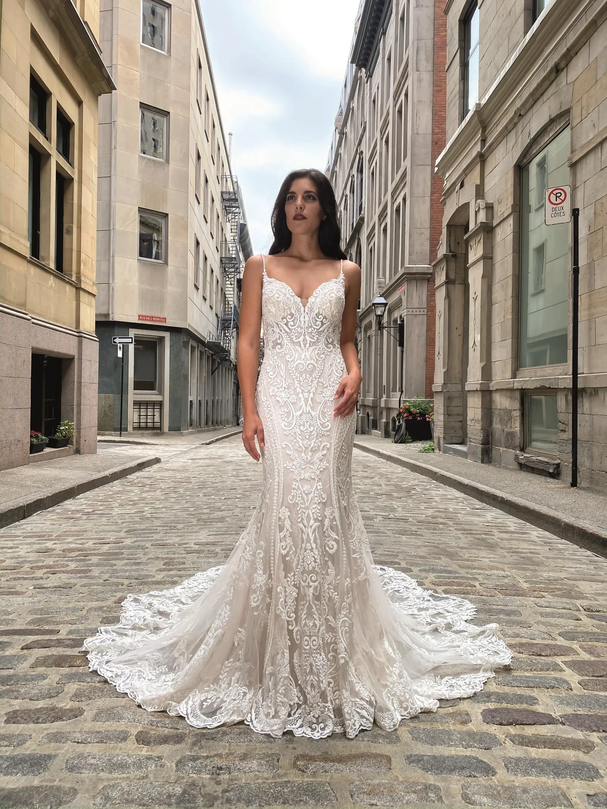 A woman poses in a flowing, intricately designed wedding dress on a cobblestone street between tall buildings.