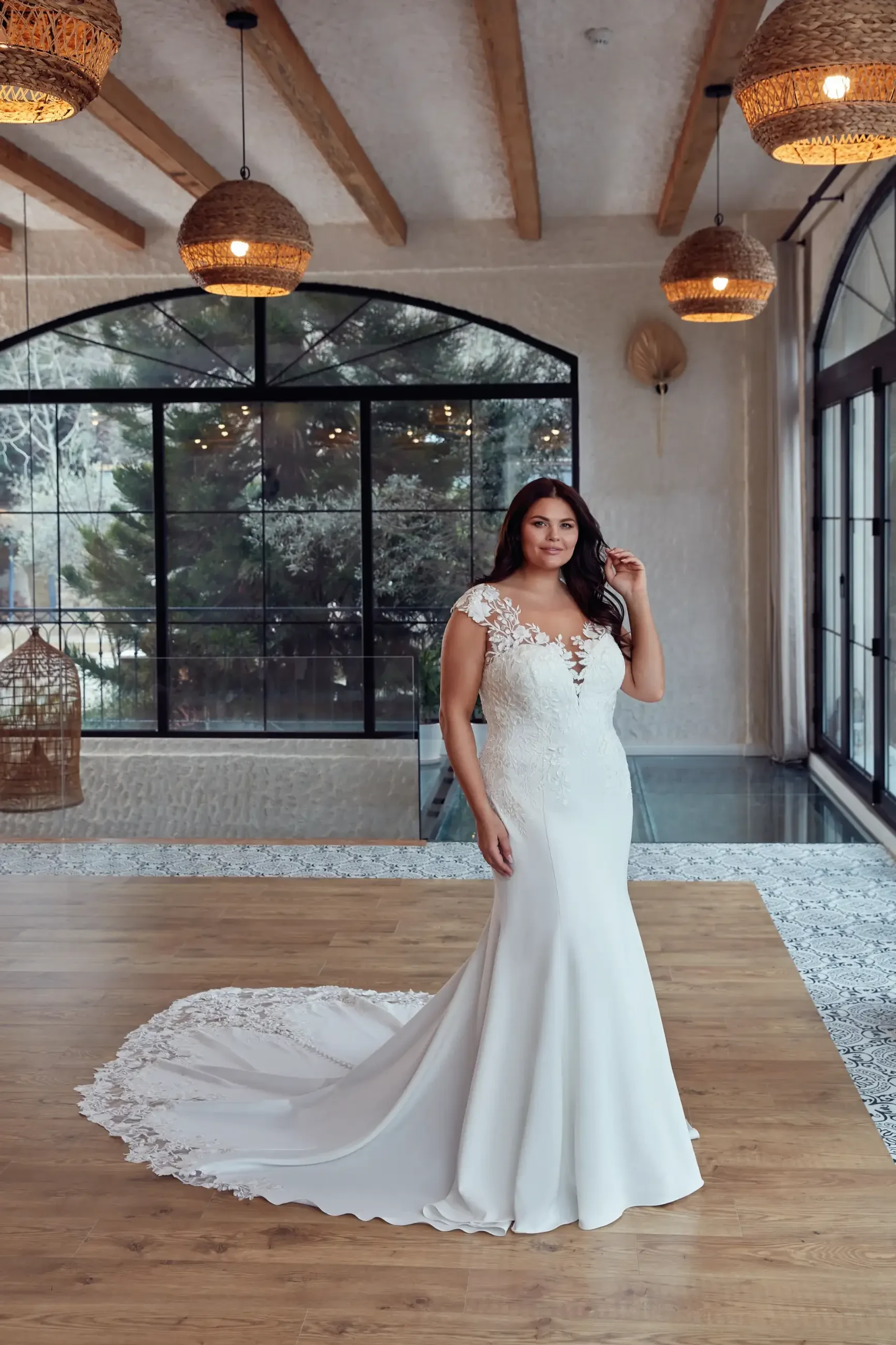 Model is wearing a plus size fit and flare wedding dress with cap sleeves and lace and a long train
